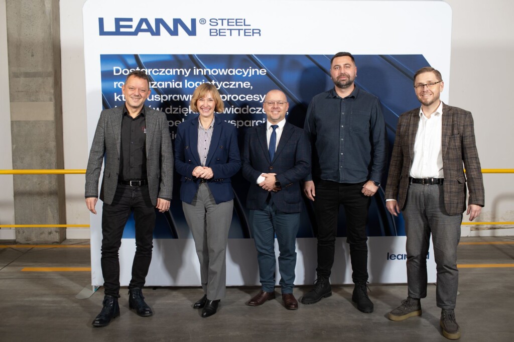 Pięć osób w formalnych strojach stoi przy tle z logo i hasłem firmy LEANN, w hali produkcyjnej