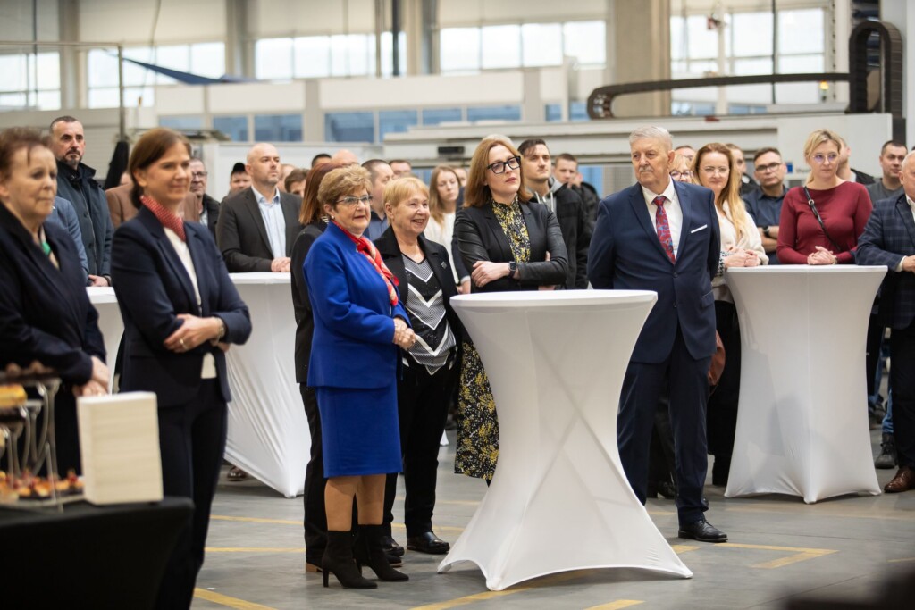 Grupa osób w eleganckich strojach, słuchających przemówienia w zakładzie produkcyjnym z białymi stołami koktajlowymi