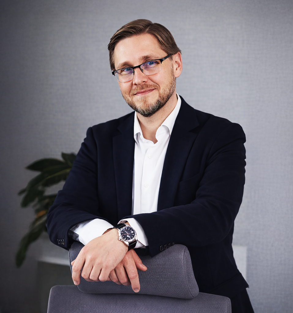 Mężczyzna w okularach, w garniturze, opiera się na fotelu w nowoczesnym biurze z rośliną w tle. Na zdjęciu prezes Łukasz Stańczyk.