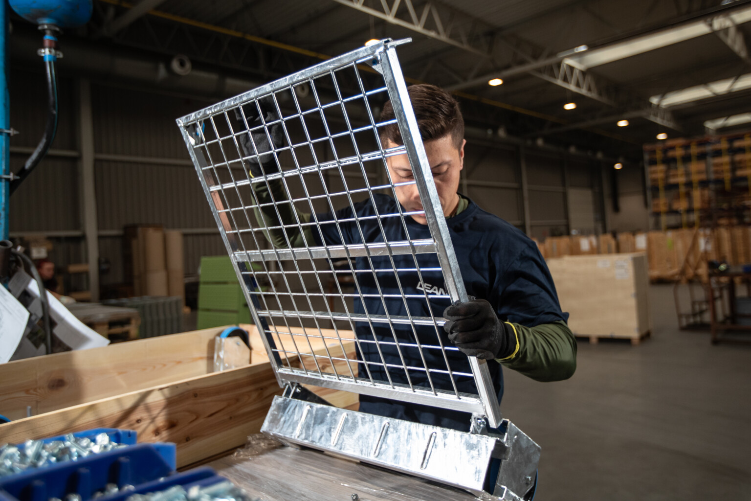 Mężczyzna w roboczej koszulce montuje metalową siatkę w hali produkcyjnej z drewnianymi pudełkami w tle