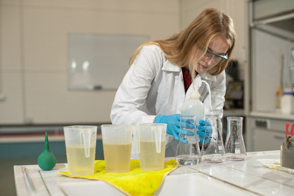 Młoda kobieta w laboratorium napełnia próbówki różnymi cieczyami, ubrana w biały fartuch i rękawiczki ochronne