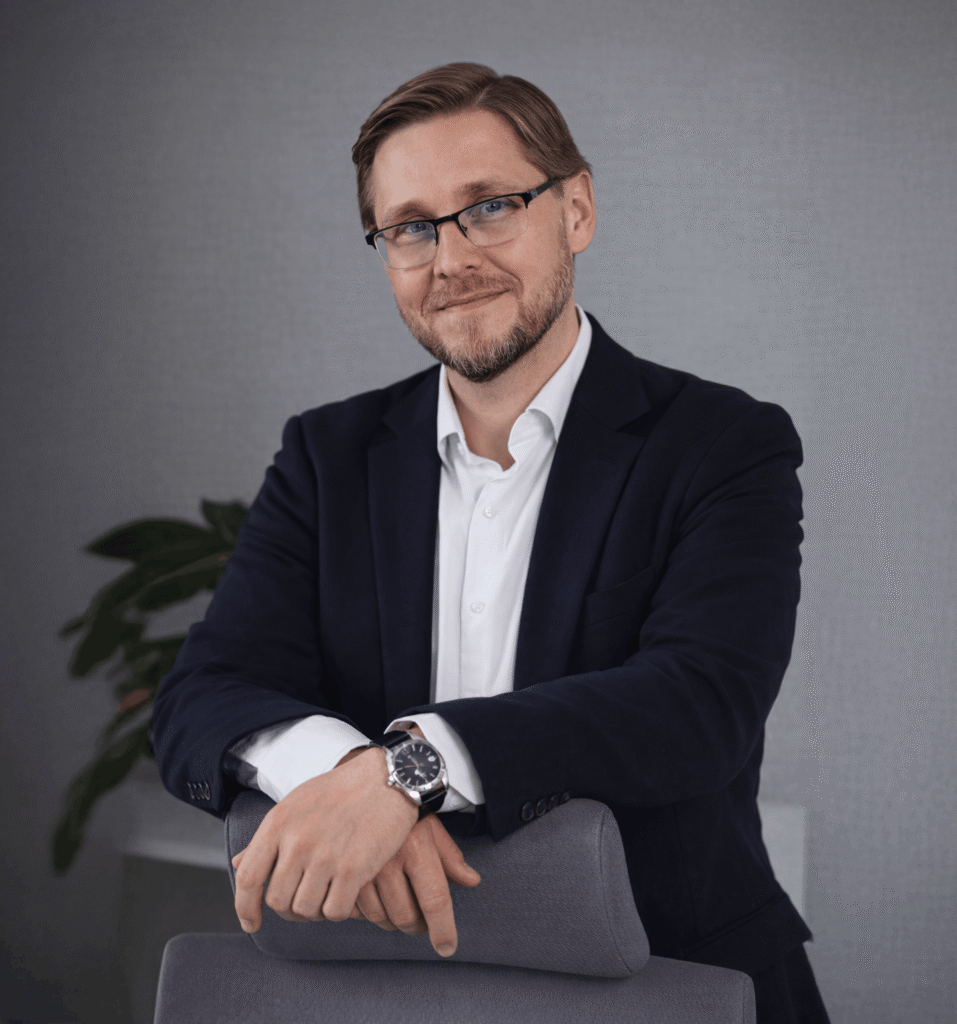 Mężczyzna w garniturze z okularami, opierający się na krześle w nowoczesnym biurze, z zielonymi roślinami w tle. Na zdjęciu prezes Łukasz Stańczyk.